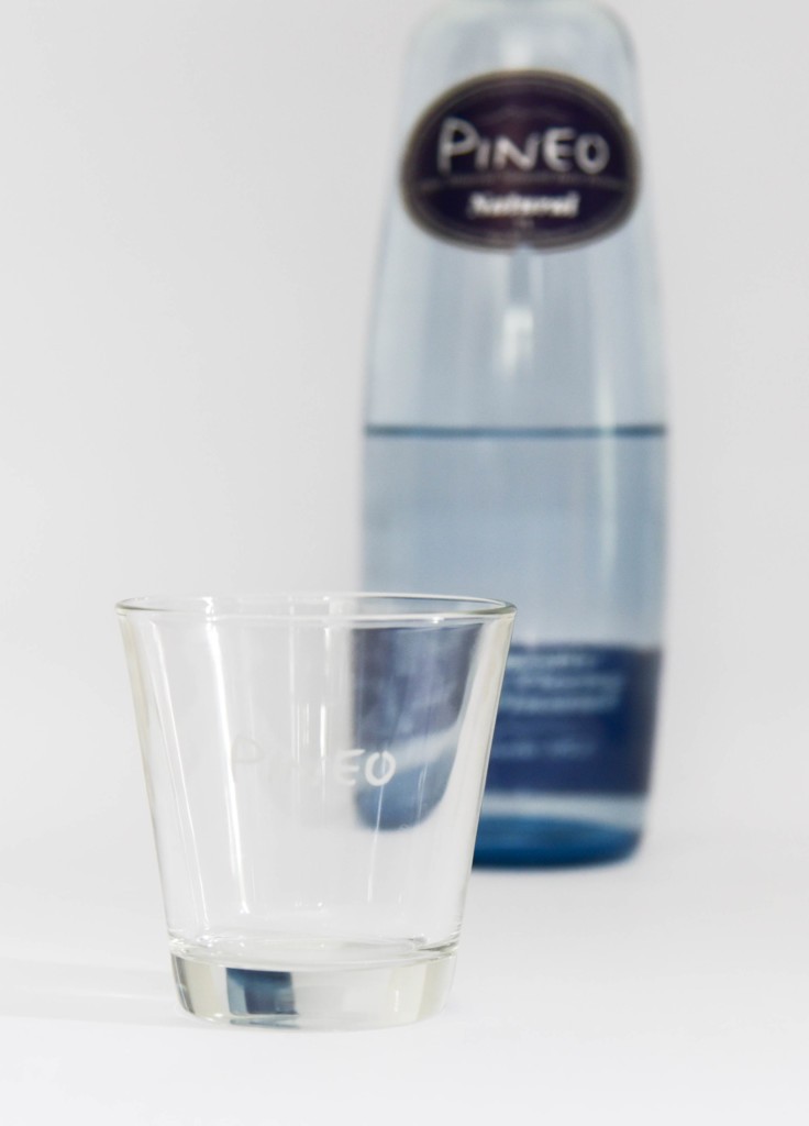 Pineo Glasflasche mit Glas im Vordergrund. Natürliches, selbst entspringendes Mineralwasser aus den spanischen Pyrenäen.
